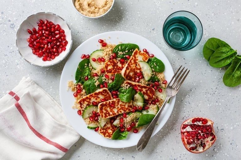 Overhead view of a colorful salad with pomegranate seeds, greens, and nuts on a white plate with water glass and pomegranate